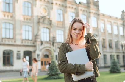 Smiling student holding a laptop outdoors in front of a historic university building.